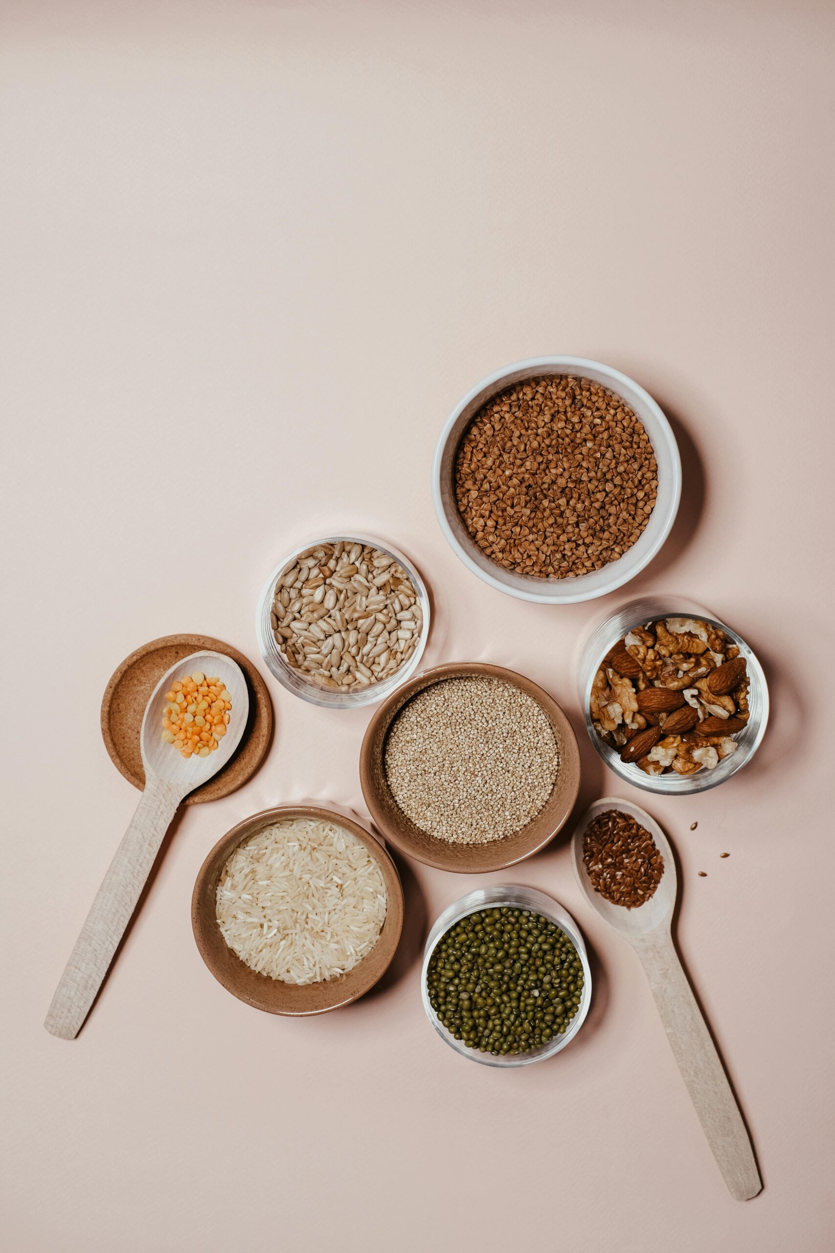 Flat lay of various grains and nuts in bowls and wooden spoons on neutral background.